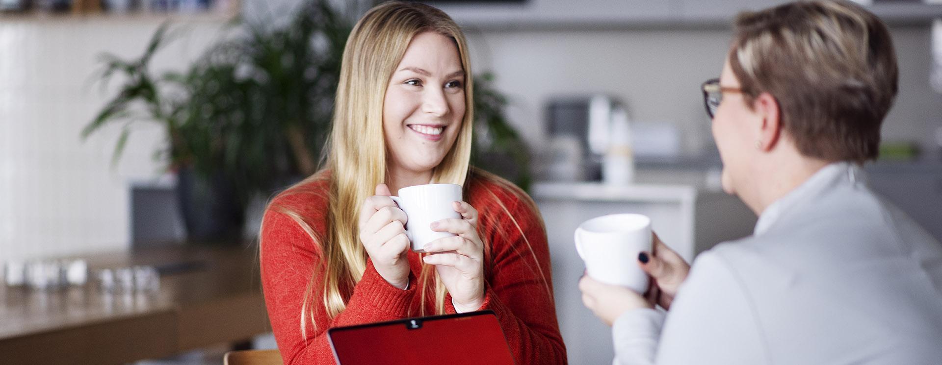 Två kvinnor sitter med varsin kaffekopp i handen och ler mot varandra.