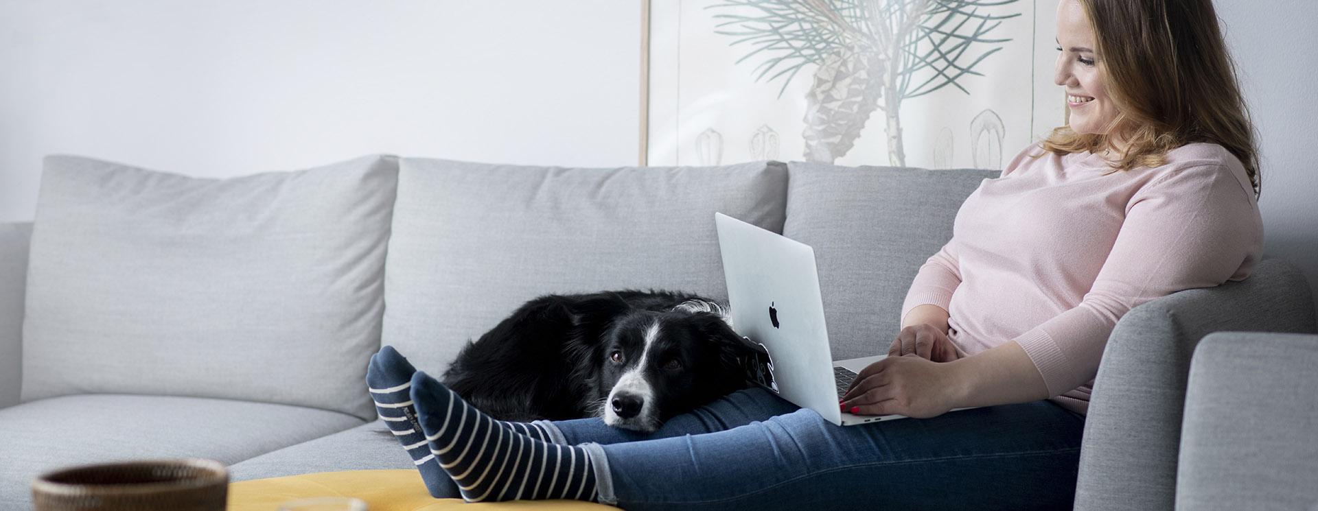 En kvinna sitter i en soffa med fötterna på bordet. I knät har hon en laptop och en svart, stor hund som vilar med huvudet på hennes ben.